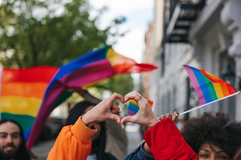 Genderqueer and non-binary friends making a heart shape against a rainbow flag