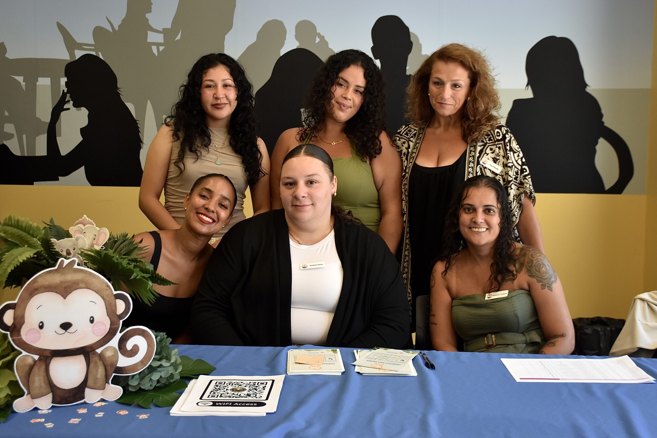 Six women pose together at a table decorated with a jungle animal theme, including a cardboard cutout of a monkey and plants, at the New Bedford FRC Community Baby Shower. Materials on the table include a WiFi access sign, a QR code, and event flyers.