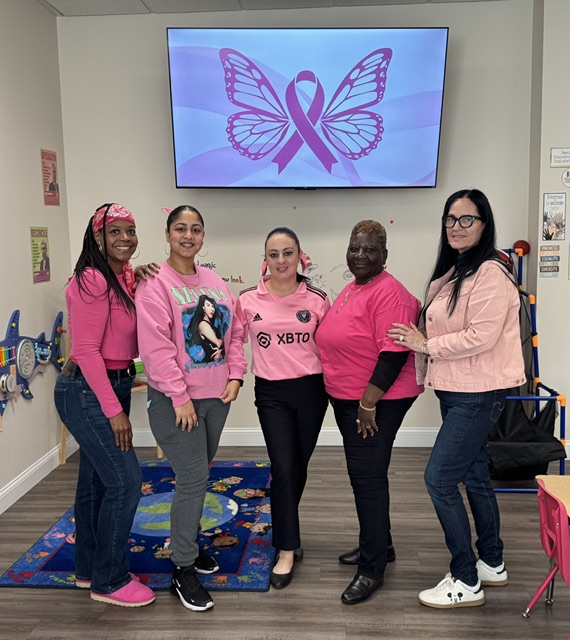 Five Springfield FRC staff members stand together dressed in pink in a classroom space. A TV behind them shows a purple butterfly with a pink awareness ribbon.